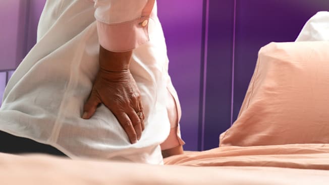 close up of person sitting on edge of bed with hand on their hip, peach colored sheets and pillows in the background