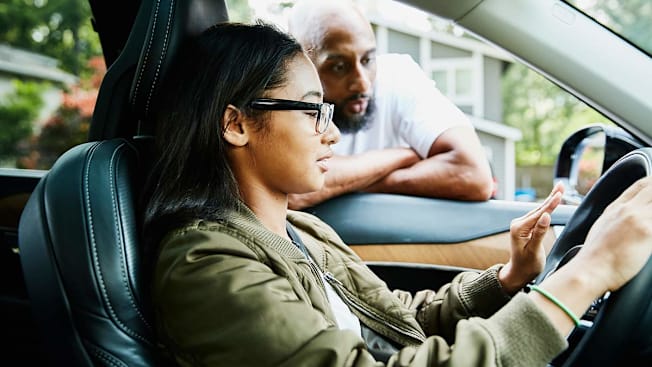 A teen sitting in the driver's seat of a car with their father leaning into the driver's side window.