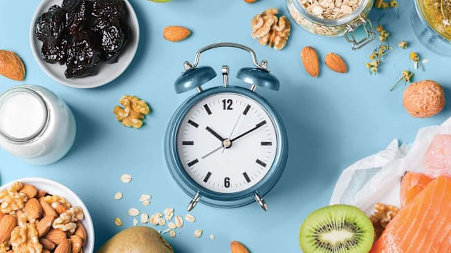 overhead view of clock with walnuts, almonds, kiwi, oatmeal, milk, prunes, and salmon around it