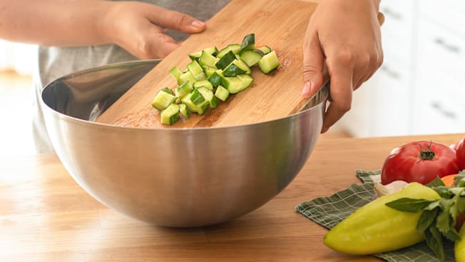 Person sliding cucumbers from wooden cutting board to metal bowl in kitchen