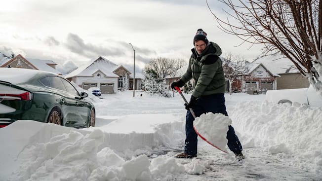 person shoveling deep snow from driveway in suburban neighborhood with other houses in background