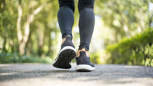 Closeup of the back of a person's shins and sneakers as they walk down a tree-lined paved road.