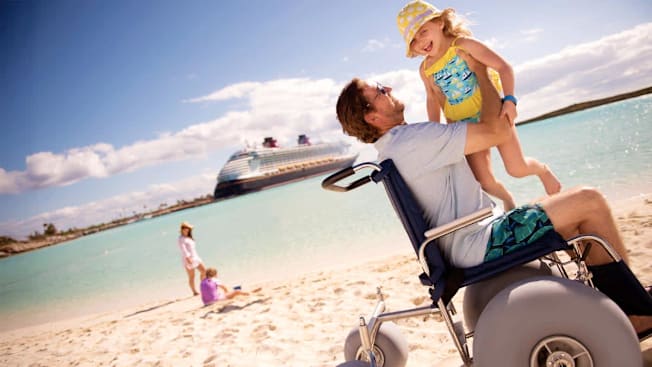 A man seated in a beach-accessible wheelchair lifts a smiling young girl wearing a yellow polka-dot sunhat and a yellow-and-blue swimsuit. They are on a sandy beach at Disney Castaway Cay with turquoise water and a Disney Cruise Line ship in the background. A woman and another child play in the sand behind them.