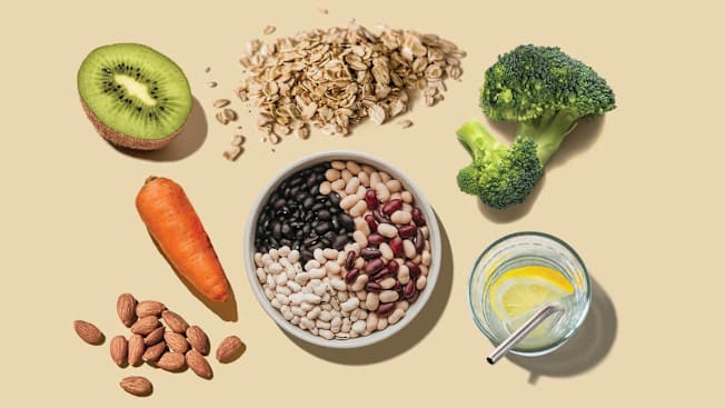 half a kiwi, carrot, almonds, glass of water with metal straw, broccoli floret, oatmeal pile, and bowl of beans on beige background