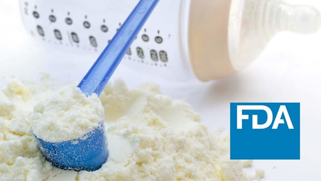 A close-up of powdered infant formula with a blue plastic scoop partially buried in the powder, a baby bottle visible in the background, and an FDA logo displayed on the right side.