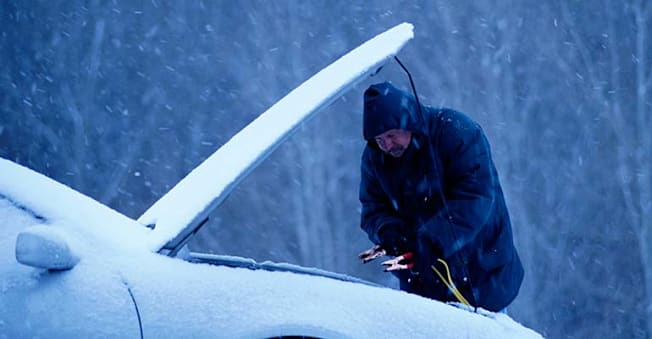Person using jumper cables on a car during a snow storm.