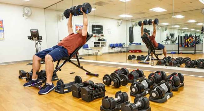 tester in gym lifting adjustable dumbbells during testing with other dumbbells next to weight bench