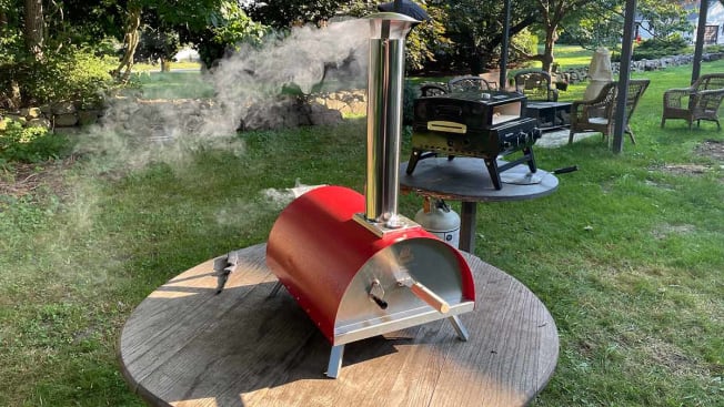 A LePeppe pizza oven in the foreground with smoke billowing out of its chimney and a Bakerstone pizza oven in the background.
