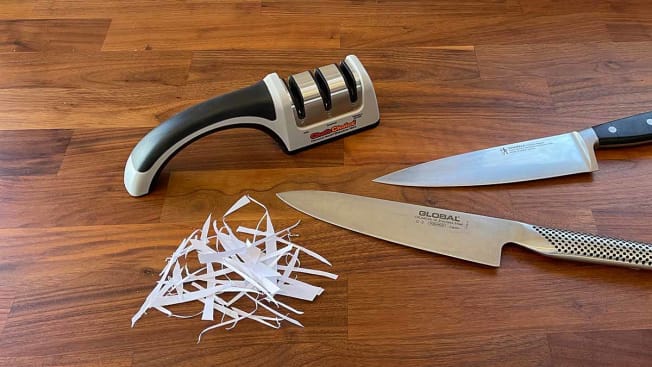 Chef's Choice manual knife sharpener seen on a cutting board with two sharp knives and cut strips of paper.