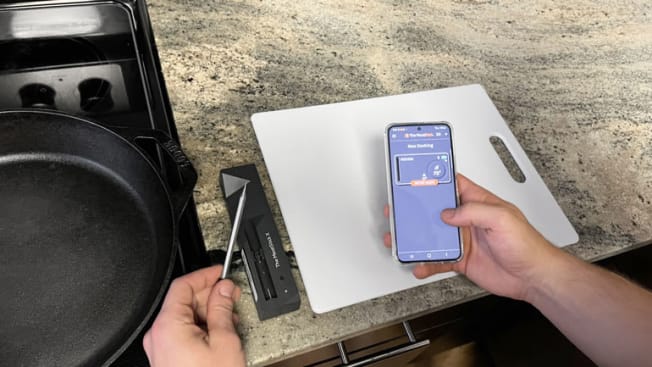 detail of person stand in front of stove and countertop holding The MeatStick X meat thermometer in one hand and smartphone with The MeatStick X app open in the other hand