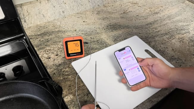 detail of person stand in front of stove and countertop holding ThermaPro meat thermometer in one hand and smartphone with ThermaPro app open in the other hand