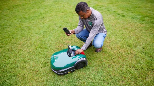 Consumer Reports tester surrounded by lawn- holding smartphone while inspecting Robomow robotic mower