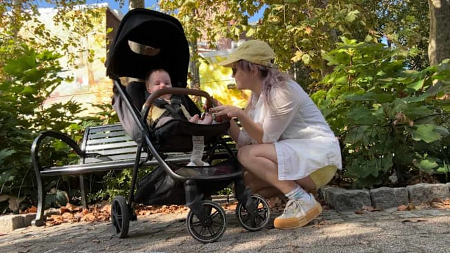baby in Nuna TRVL LX stroller with parent squatting down in front of stroller looking at baby, in park in front of a park bench
