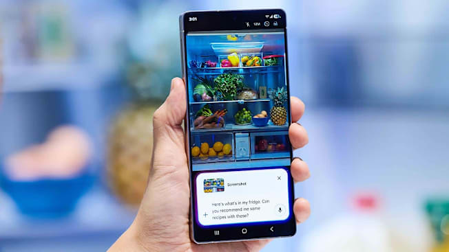 A hand holding the Samsung S25 Ultra with an example of Gemini AI being used on its screen of a prompt asking what recipe can be made from the contents of a refrigerator.