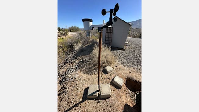 Ambient Weather Weather Station set up on wooden pole with concrete slabs with desert landscape, building, and mountains in background