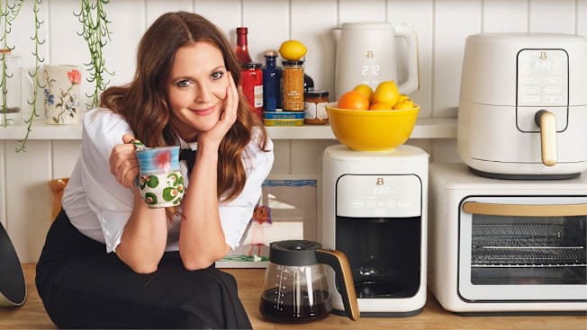 Drew Barrymore sitting on a countertop surrounded by her brand's appliances with a cup in her hand