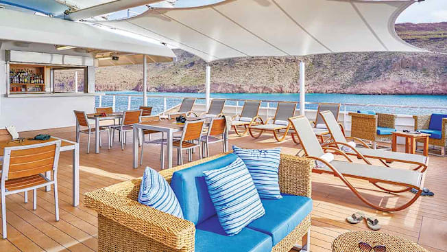 The National Geographic cruise ship's sun deck featuring a bar, dining tables, and lounge chairs with ocean and coastal views.