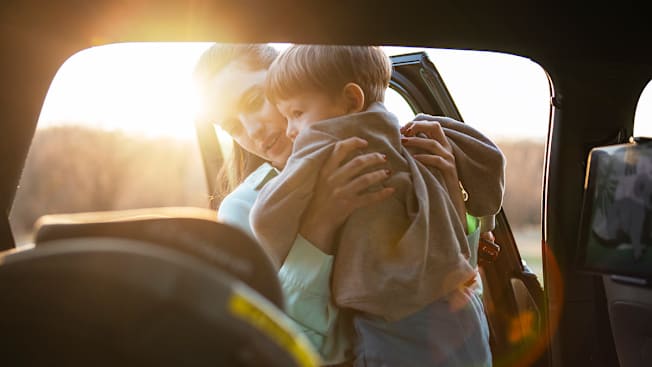 A mother gently lifts her young child out of a car seat in the back of a vehicle during a bright, sunny day.