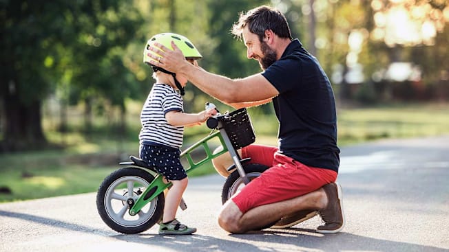 A father kneels on a paved path to adjust a bright yellow helmet on his young child, who is sitting on a green balance bike.