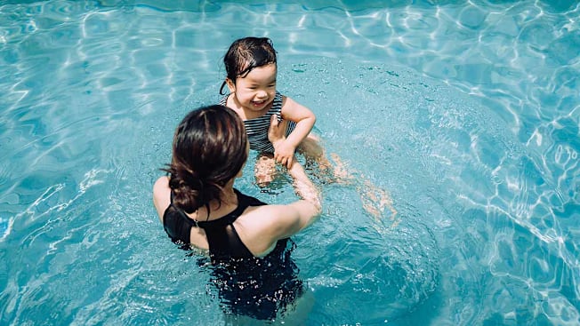 An overhead view of a mother smiling and holding her toddler as they play together in a clear, blue swimming pool.