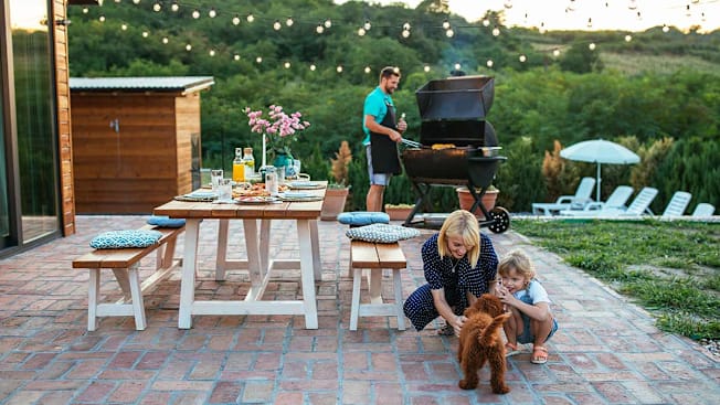 An outdoor patio scene where a man cooks at a large grill while a woman and a young girl crouch nearby to pet a small brown dog.
