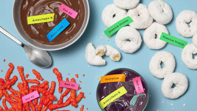 Chocolate pudding, powdered mini donuts, spicy snack chips, and purple soda displaying food additives including Red 40, aspartame, sucralose, acesulfame potassium, and titanium dioxide.