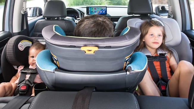 Three car seats are installed side-by-side in the backseat of a car, seen from the perspective of the trunk looking forward. On the left, a baby sits in a rear-facing infant car seat; in the middle, a young boy sits in a rear-facing car seat; and on the right, a young girl sits in a forward-facing car seat looking forward.