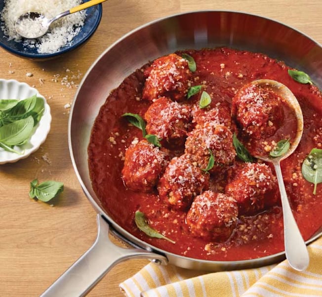 A stainless skillet of meatballs and marinara sauce, garnished with fresh basil leaves and grated parmesan cheese.