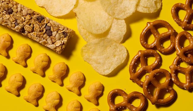 Lay's Classic Potato Chips, Quaker Chewy Bars Chocolate Chip, Snyder's of Hanover Mini Pretzels, and Pepperidge Farm Goldfish Cheddar Crackers arranged on a bright yellow background.