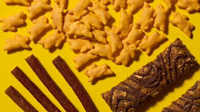 Annie's Organic Cheddar Bunnies, Slim Jim Mild Snack Size Sticks, and Clif Zbar Chocolate Chip bars arranged on a bright yellow background.