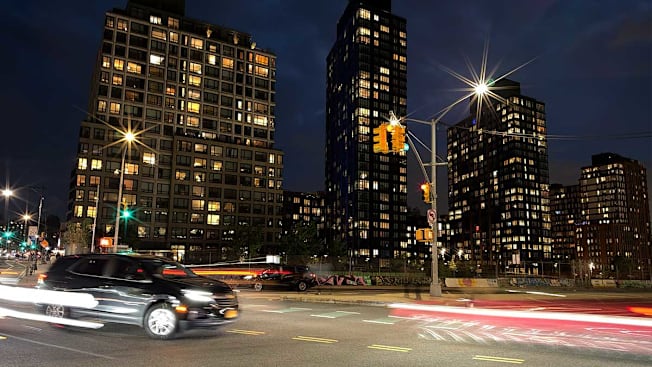 A nighttime urban street scene showing tall residential high-rises with lit windows against a dark blue sky. In the foreground, a dark SUV drives across an intersection, leaving motion-blurred light trails from passing car headlights and taillights.