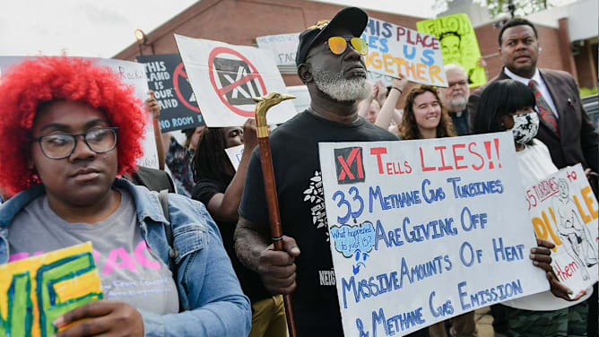 A group of community members in Memphis, TN participates in an outdoor protest. In the center, an older man with a gray beard and orange-tinted sunglasses holds a walking cane and a large hand-painted sign that reads, "[xAI Logo] Tells Lies!! 33 Methane Gas Turbines are giving off massive amounts of heat & Methane Gas Emission." To his left, a young person with vibrant red hair and glasses looks toward the side. In the background, other protesters hold signs, including one that says "This Place Ties Us Together." The scene depicts a grassroots environmental demonstration against a local industrial project.