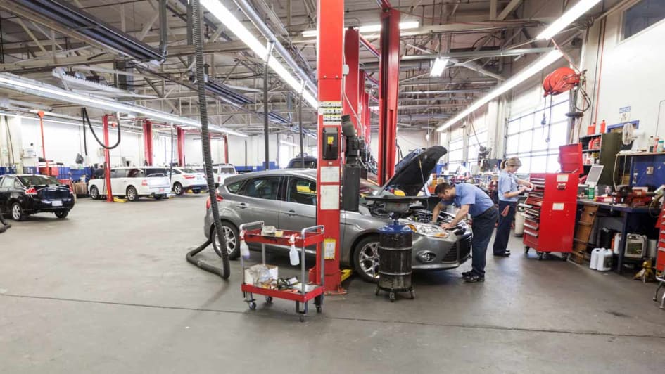 auto repair shop with several cars and two people working, one person looking under hood of car