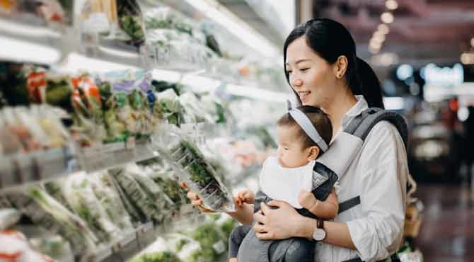 An image of an Asian woman checking a food package's label while holding a baby in a supermarket.